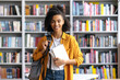 © Kateryna - Portrait of an african american happy pretty, smart confident female student with backpack, stylishly dressed, holding books, stands in the university library, looks at the camera, smiling friendly