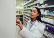 © StratfordProductions - Young woman pharmacist wearing labcoat searching for medicine in shelf in chemist
