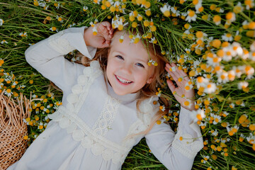 happy little girl in a cotton dress lies in a field of daisies in the summer at sunset. laughs, view from above