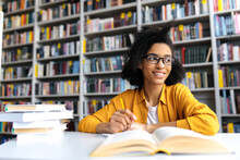 Teenage African American Female Pretty Student With Glasses, Studying While Sits At The Table In A College Library, Reads Books To Searching Information For A Lesson Or Exam, Doing Homework,looks Away