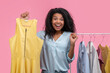 Studio portrait of excited smiling african american girl holding hanger with illuminating yellow dress in hand while standing near the rack of pastel clothes in showroom