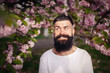 © endrews21 - Cheerful stylish man with a long dark beard and mustache in a white T-shirt near blooming sakura trees in the park in nature. Adult guy with gray hair and dyed hair