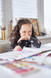 © boytsov - Laughing kid girl sitting at the table in headphones doing homework drawing using pencils or crayons. Homeschooling or online education concept. Vertical image. High quality photo