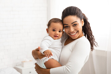 joyful black mom holding and hugging adorable baby in bedroom