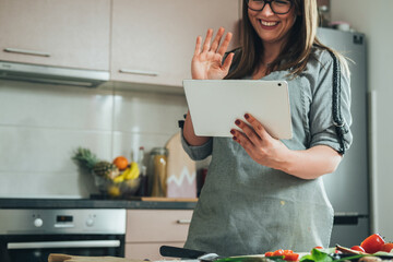 Cheerful woman making video call from digital tablet while cooking lunch in kitchen. Smiling housewife waving and saying hello or goodbye on online video call, working at home concept