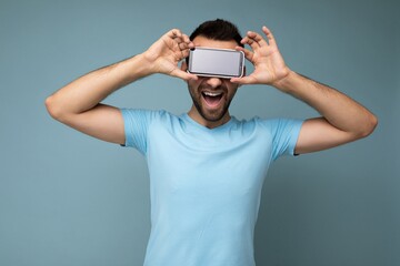 Positive smiling handsome young unshaven brunet man with beard wearing everyday blue t-shirt isolated over blue background holding and showing mobile phone with empty display for mockup