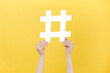 Cropped images of female hands holding large big white hashtag sign, viral web content, internet promotion, isolated on yellow studio background wall. Concept of trendy social media posts and blogging
