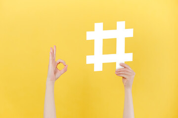 Close up of unrecognizable female holding large big hashtag sign and showing okay gesture, viral web content, internet promotion, isolated on pastel yellow wall. Blogging blog website strategy concept