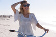 © Wavebreak Media - Smiling caucasian woman with sunglasses, white top and shorts sitting a bicycle at the beach