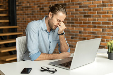 Fatigued hipster using laptop for working, a guy in smart casual wear is rubs the bridge of the nose, feels tired and burnout, sitting at the desk in modern office with eyes closed