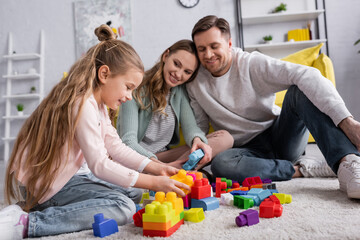 Wall Mural - Smiling kid playing building blocks on carpet near blurred parents.