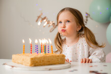 Cute Little Girl Blows Out Candles On A Birthday Cake At Home Against A Backdrop Of Balloons. Child's Birthday