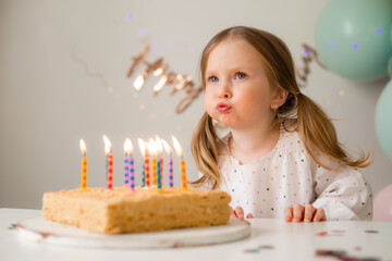 cute little girl blows out candles on a birthday cake at home against a backdrop of balloons. Child's birthday