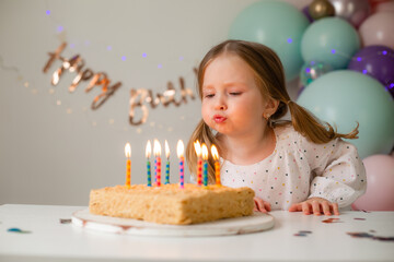 cute little girl blows out candles on a birthday cake at home against a backdrop of balloons. Child's birthday