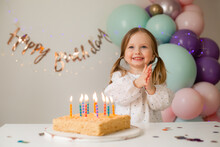 Cute Little Girl Blows Out Candles On A Birthday Cake At Home Against A Backdrop Of Balloons. Child's Birthday