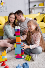 Wall Mural - Happy girl playing colorful building blocks near parents in living room.