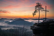Palatinate Forest on a sandstone rock with a view of the sunrise in the valley is fog and on the rock there is a cross and a lonely tree