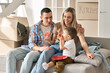 © insta_photos - Happy young family sitting on sofa with kid feeding with bread and jam. Joyful family in living room at moving day, boxes on background, mom and dad happy new householders renters tenants concept.