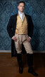 A handsome Regency gentleman wearing a gold waistcoat, breeches, and a black long jacket and standing in a room with blue wallpaper and a wooden floor