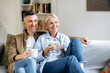 © Kateryna - Glad mature caucasian married couple in stylish casual clothes, sitting on a comfortable sofa in the living room, holding cups of tea, chatting, spending time at home, looking away, smiling