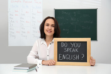 Poster - Teacher holding small chalkboard with inscription Do You Speak English? at table in classroom