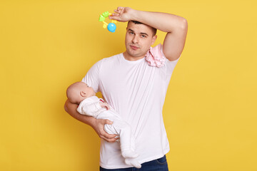 Wall Mural - Tired young father with newborn baby in hands, male taking care of his small daughter or son, keeping hand with toy on forehead, being exhausted of lots work, isolated on yellow wall.