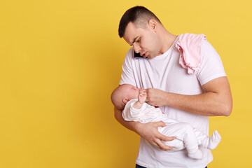 Wall Mural - Caucasian young father holding newborn baby in hid hands, calming down little kid, giving nipple, talking phone while taking care of his daughter, isolated over yellow background.