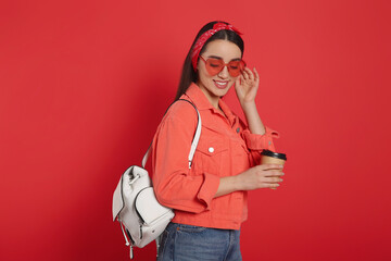 Poster - Beautiful young woman with stylish leather backpack and cup of coffee on red background