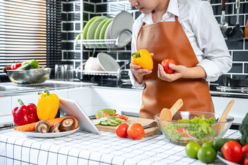 Wall Mural - Asian housewife holding bell pepper and tomato in hands during watching tutorial cooking class online on tablet