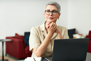 Wall Mural - Stylish mature woman with grey hair wearing glasses sitting at home in front of a laptop