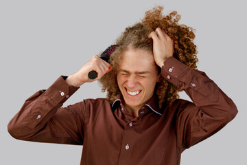 A long-haired curly-haired guy in a brown shirt on a gray background uses a metal round comb. Emotions before a haircut in a hairdresser. Pain from combing