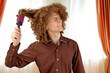 © Yuliia - A long-haired curly-haired guy in a brown shirt uses a metal round comb. Emotions before a haircut in a hairdresser. Pain from combing