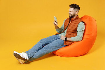 Wall Mural - Full length young smiling happy fun caucasian man 20s years old wear orange vest mint sweatshirt sitting in beanbag bag chair hold mobile cell phone chat isolated on yellow background studio portrait.