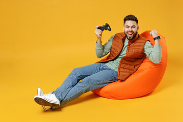 Full length young excited man 20s in orange vest mint sweatshirt sitting in beanbag bag chair playing pc game with joystick console do winner gesture clench fist isolated on yellow background studio