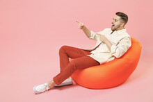 Full Length Young Happy Smiling Fashionable Man In Jacket White T-shirt Sitting In Bean Bag Chair Point Index Finger Aside On Workspace Area Mock Up Isolated On Pastel Pink Background Studio Portrait.