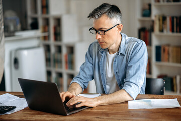 Focused smart successful gray-haired senior caucasian man wearing glasses, freelance, businessman or manager, works remotely on laptop in living room, texting with colleague or client,preparing report