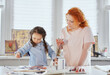 © boytsov - Family leisure activity at home concept. Mother and small daughter drawing together using paintbrushes and watercolors or acrylic. Young woman helping or teaching daughter sitting on a table to paint