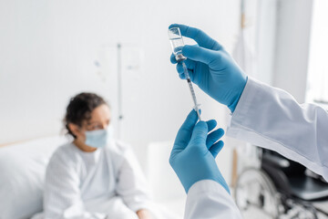 blurred african american woman in medical mask near doctor in latex gloves holding syringe and bottle with vaccine