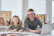 © boytsov - Cute small kid girl drawing next to dad who is working online in headphones with laptop at the table. Attractive family busy looking straight to camera happy smiling during covid-19 lockdown