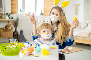 Beautiful happy son and mother in headbands with bunny ears decorating eggs using paintbrush and bright colors of paint, preparing to celebrate Easter holiday, having fun spending time together at hom