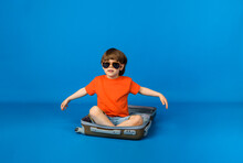 A Little Boy Traveler Wearing Sunglasses Sits In A Suitcase On A Blue Background With Space For Text