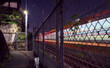 Long-exposure capture of light trails made by a train passing by an alleyway, separated from the tracks by a chain link fence