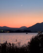 A crescent moon rising over Lake Kawaguchi and a blue and orange sky at dusk, with low mountains and a boat visible