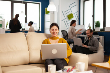 Poster - Businesswoman sitting on couch holding laptop, smiling at camera while diverse colleagues working in background. Multiethnic coworkers talking about start up financial company in modern business