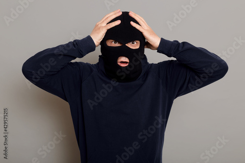 Shocked and frightened criminal, thief catching on place of committing crime, looking at camera with frightened eyes, holding his head, does not understand what to do, isolated over gray background.