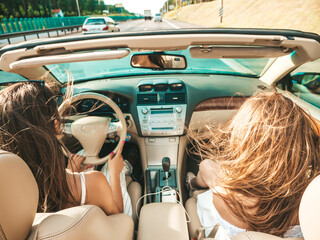 Portrait of two young beautiful and smiling hipster female in convertible car. Sexy carefree women driving cabriolet. Positive models riding and having fun in sunglasses outdoors. Enjoying summer days