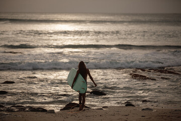 Poster - Young female surfer walking towards the ocean waves carrying a board. Surfing at sunset