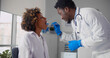 © nimito - Afro-american male doctor examining young woman throat in office.