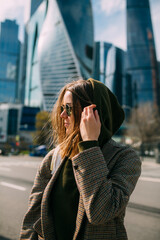 Wall Mural - A blonde young girl with a white backpack dressed in a jacket stands against the background of skyscrapers. Large windows of skyscrapers reflect the sun. The girl in the hood travels 