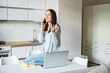 © Vadim Pastuh - Charming young woman using laptop computer for remote work from home, talks on the smartphone leaned on the table in relaxed pose, female freelancer has pleasant phone conversation